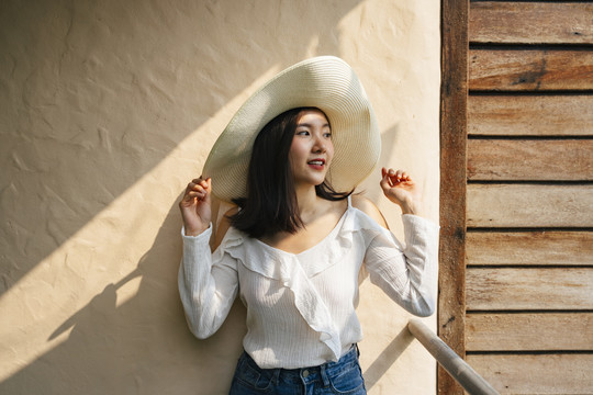 美丽的亚洲女人穿着白衬衫，戴着大帽子站在墙上。