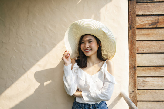 美丽的亚洲女人穿着白衬衫，戴着大帽子站在墙上。