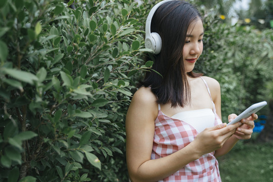身着粉色连衣裙的泰国亚裔美女站在绿叶树旁，用智能手机的无线耳机听音乐。