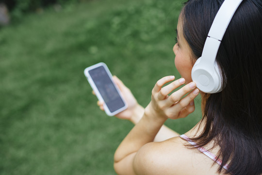 肩顶视图-阿西娜女孩用白色无线耳机听音乐，手持智能手机控制音乐平台。