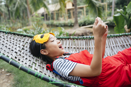 戴着卡通狮子面具的泰国小女孩躺在婴儿床上玩着表情幸福的智能手机。