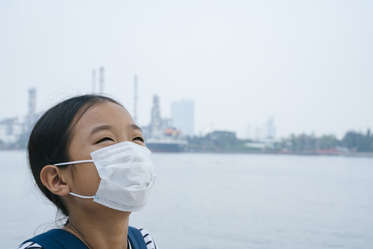 病毒防护理念——戴着防护面具的快乐亚洲小女孩，在雾霾天站在河边。到处都是污染。她不知道PM2.5有多
