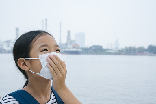 病毒防护概念-戴着防护面具的亚洲小女孩，在雾霾天站在河边。到处都是污染。用手捂住嘴。