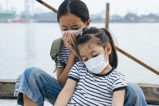 病毒防护概念-戴着防护面具的亚洲小女孩，在雾霾天站在河边。到处都是污染。两个妹妹因为PM2.5污染而