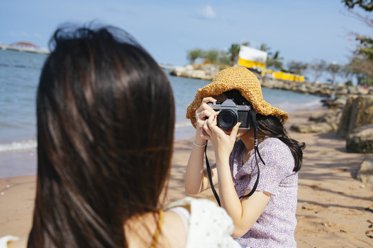 一个戴着棕色帽子的女孩给她的朋友拍了一张照片，而他们坐在海边的海滩上，在夏天的多风的日子里。
