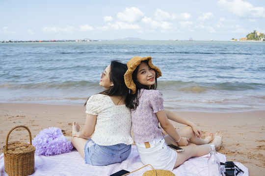 两个女孩坐在沙滩上的垫子上，拿着木篮在海边野餐。