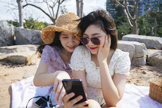 两个女孩坐在海边沙滩上的垫子上，用智能手机看照片。