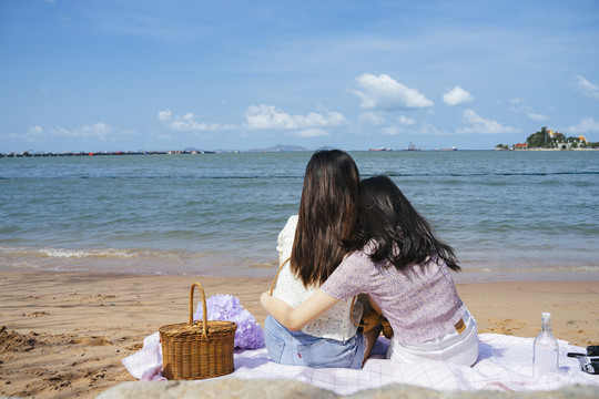 两个女孩坐在海边沙滩上的垫子上，其中一个依偎着另一个。