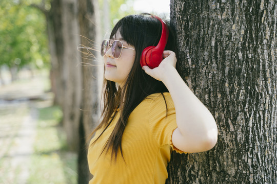 戴着红色耳机的长发女孩倚靠在大树上，在风和日丽的日子里欣赏音乐。
