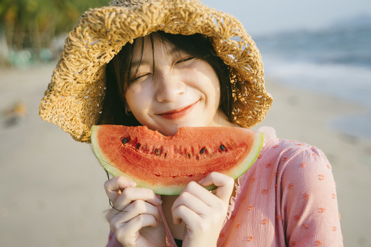 戴棕色帽子的女孩在沙滩上咬西瓜，笑了。
