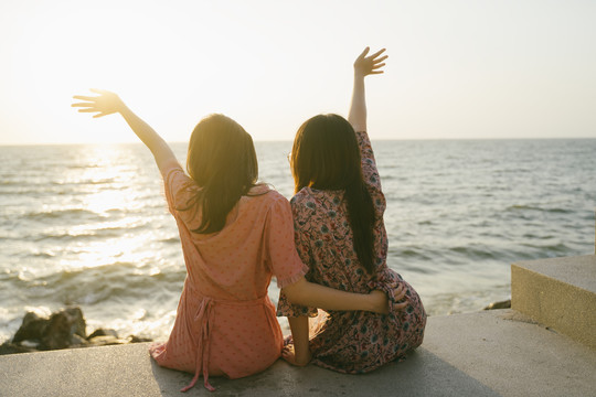 两个长发女孩坐在码头上看着夕阳，向天空举手，回击。