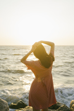 长发女孩站在海边的岩石上，悠闲地看着夕阳。