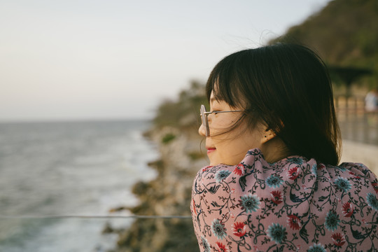 戴着墨镜的长发女孩站在海边楼台的眼镜路障旁，傍晚眺望大海。