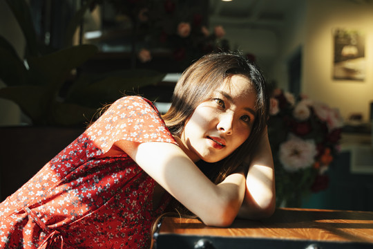 阳光下，身穿红色连衣裙的泰国亚裔年轻女子躺在屋内的桌子上。