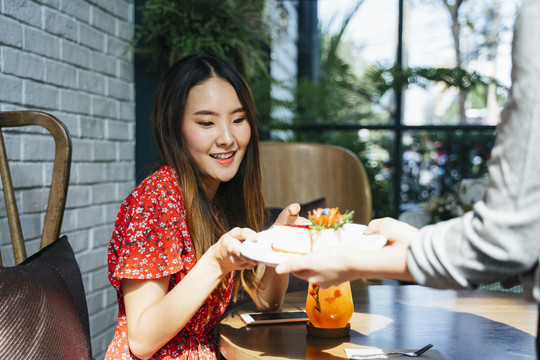 一位身穿红色连衣裙的泰国亚裔年轻女子在咖啡馆从女服务员那里拿蛋糕。