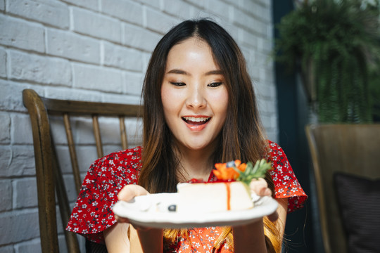 身穿红色连衣裙的泰国亚裔年轻女子，手里拿着白色盘子，盘子上放着奶酪蛋糕。快乐的表情。