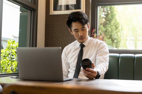 亚泰商人穿着白衬衫，正装，看着一杯咖啡在笔记本电脑上工作。
