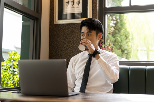 穿着白领衬衫的亚泰商人在咖啡馆喝咖啡，在笔记本电脑上工作。