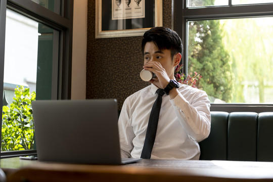 穿着白领衬衫的亚泰商人在咖啡馆喝咖啡，在笔记本电脑上工作。