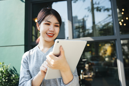 美丽的亚洲年轻女子在街上的咖啡馆背景上使用平板电脑。
