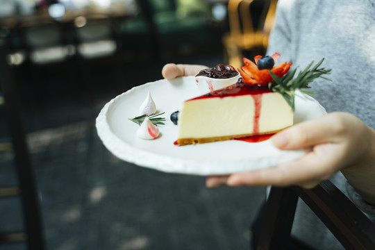 服务员的手拿着一个盘子，上面放着一片芝士蛋糕和蓝莓。