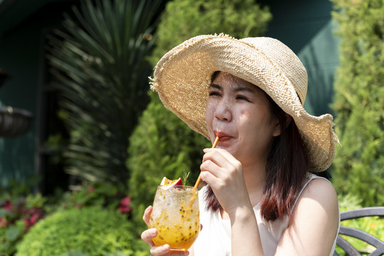 美丽的亚泰女子戴着帽子，在花园里畅饮百香果汁。