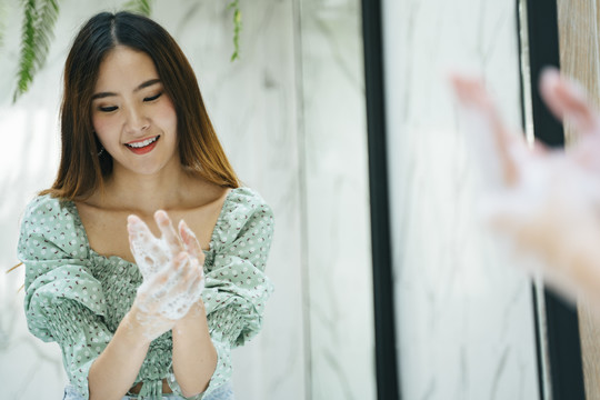 穿着时尚绿衬衫的亚洲美女在卫生间里用水和肥皂泡洗手。看着镜子。
