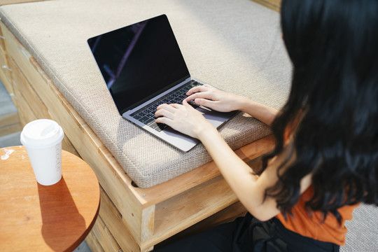 性感的亚洲女人穿着橙色衬衫在咖啡馆的笔记本电脑键盘上打字。