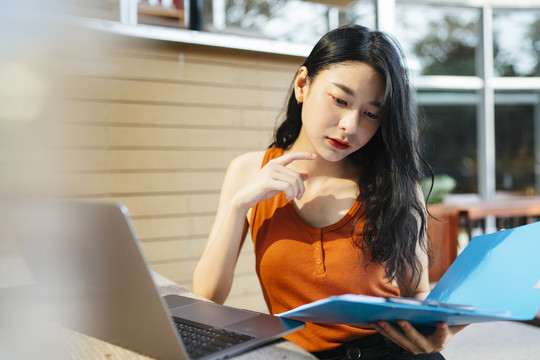 性感的亚洲女人穿着橙色衬衫在家里工作拿着蓝色的剪贴板文件看着笔记本电脑。