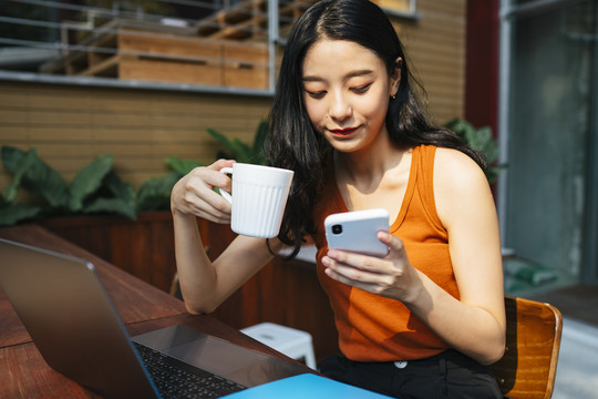 一位穿着橙色衬衫的亚洲美女在城里的咖啡馆里用笔记本电脑工作。