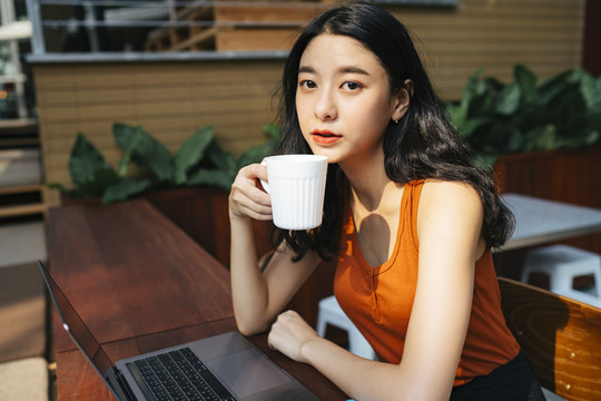 一位穿着橙色衬衫的亚洲美女在城里的咖啡馆里用笔记本电脑工作。