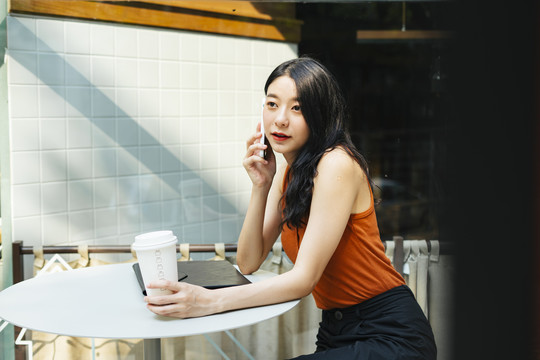 穿着橙色衬衫的泰国亚裔美女在户外咖啡厅用智能手机聊天。
