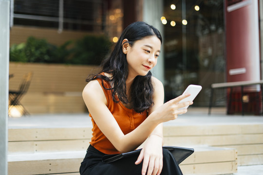 亚洲美女穿着橘色衬衫用智能手机，坐在城市的楼梯上。