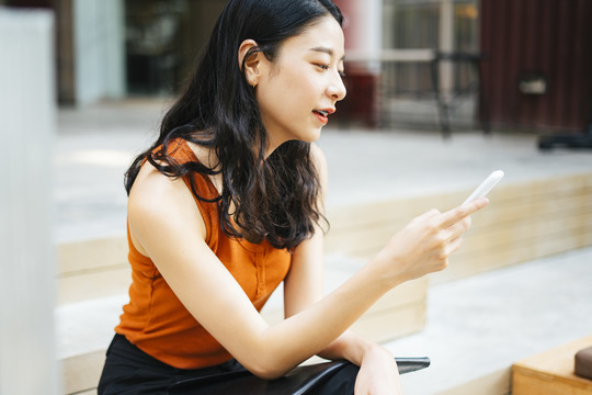 亚洲美女穿着橘色衬衫用智能手机，坐在城市的楼梯上。