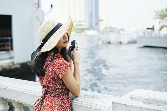 一名泰国亚裔旅行家，身穿红色连衣裙，头戴帽子，手持智能手机在河边。
