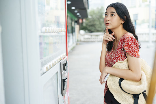 年轻漂亮的泰国亚裔旅行家，穿着红色连衣裙，从投币机上购买商品。