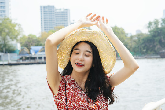 年轻漂亮的泰国亚裔旅行家，身穿红色连衣裙，戴着帽子，闭着眼睛，在河边码头举手。