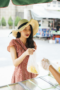 年轻漂亮的泰国亚裔旅行家，穿着红裙子，戴着红帽子，从街边小摊上买水果。