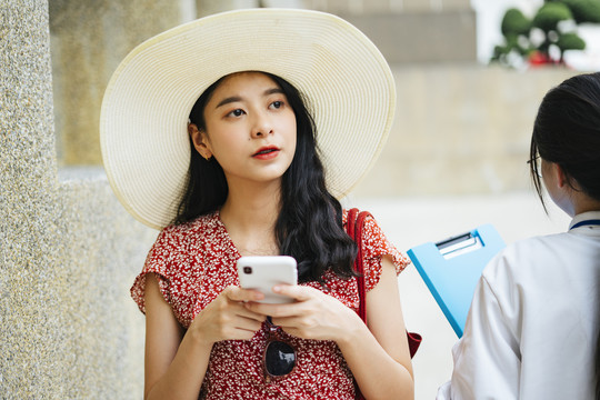 美丽的亚洲女子身穿红色连衣裙，戴着帽子，手持智能手机，神情茫然地走进城市，走进导游工作人员。
