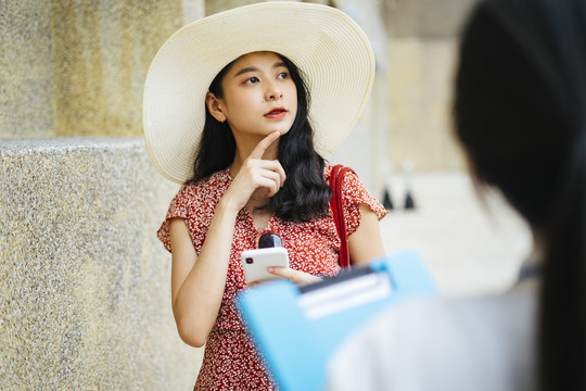 美丽的亚洲女子身穿红色连衣裙，戴着帽子，手持智能手机，神情茫然地走进城市，走进导游工作人员。
