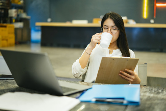 放松在办公室工作的女职员从杯子里喝咖啡，在笔记本电脑前读回形针上的信息。