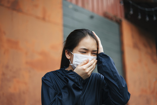 一名戴口罩的亚洲妇女因城市烟雾而咳嗽。双手捂着头。不健康的状况。