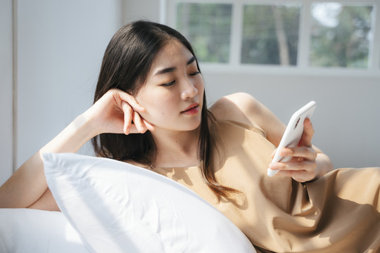 一位长发女士弯腰躺在枕头上，用智能手机查看自己的社交媒体账户。