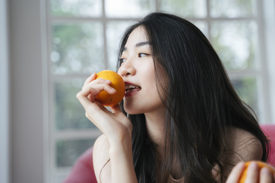 活泼的长发女孩坐在粉色沙发上，手里拿着橘子，咬着橘子。