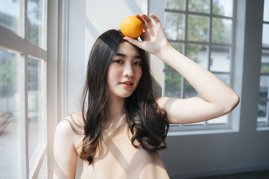 早上，一个长发卷曲的女孩站在窗前，手里拿着橘子，把橘子放在头上。