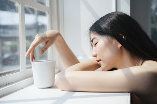 长发女孩坐在窗前，在晨光下端着一杯咖啡。