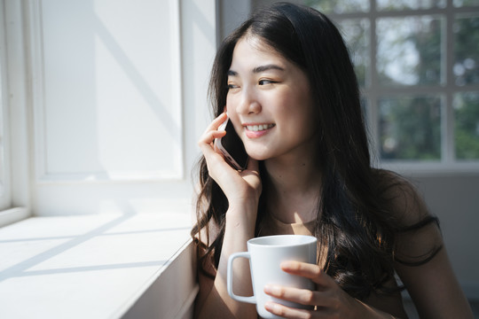 卷曲的长发女孩坐在窗前拿着热巧克力和朋友通电话。