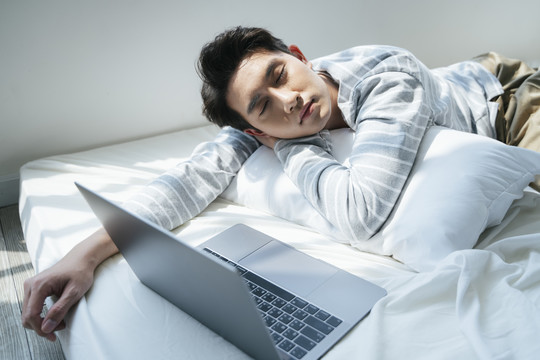 一名男子躺在白色的床上，而他在家工作时由于公司的政策疾病爆发，在视频会议后打个盹。