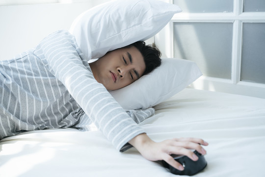 一个男人睡在白色的床上，早上闹钟把他叫醒，他用枕头捂住耳朵。