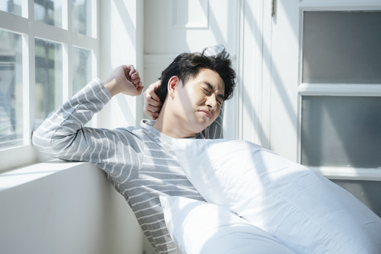 一名男子在晨光下坐在靠窗的床上，舒展身体缓解疼痛。